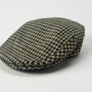 May include: A flat cap featuring a classic houndstooth pattern in shades of dark green and off-white. The cap has a rounded, structured crown and a short, stiff brim that curves slightly, showcasing a woven fabric texture throughout the exterior.