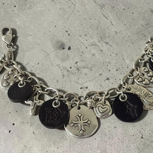 May include: Silver charm bracelet with a variety of charms, including hearts, crosses, and black circular charms. The bracelet has a lobster clasp and is shown on a textured surface. The charms feature detailed designs.