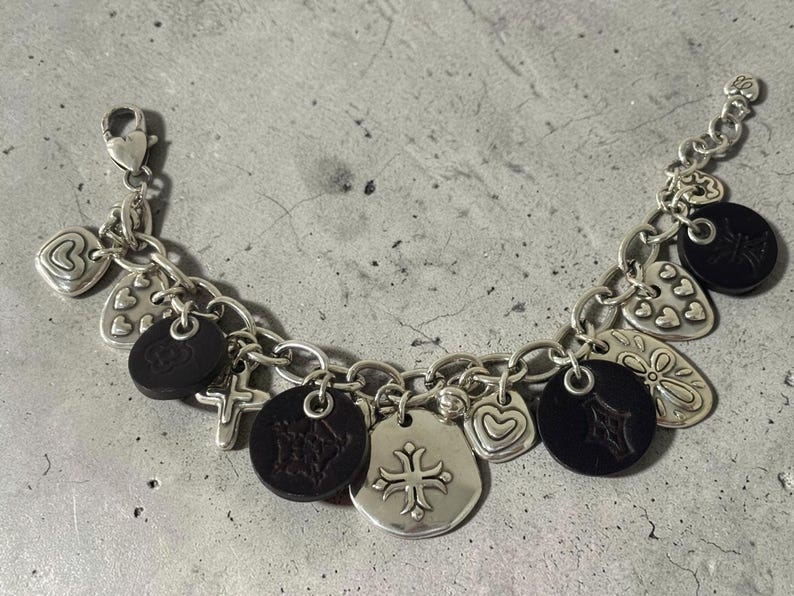 May include: Silver charm bracelet with a variety of charms, including hearts, crosses, and black circular charms. The bracelet has a lobster clasp and is shown on a textured surface. The charms feature detailed designs.
