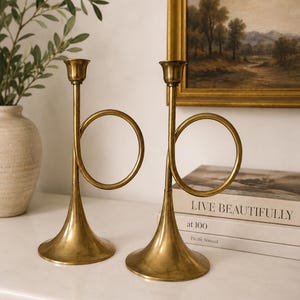 Puede incluir: Un par de candelabros de latón con forma de trompa de postillón, cada uno con un bucle circular, un tallo vertical largo, una base acampanada y una pequeña copa en la parte superior para sostener una vela. El metal tiene un acabado dorado pulido.