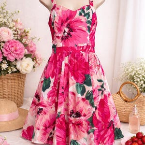 May include: A bright pink floral print dress with a sweetheart neckline and spaghetti straps. The dress has a fitted bodice and a flared skirt, displayed on a mannequin. Accessories include a straw hat, sunglasses, and a basket of flowers. The dress is a summer style.