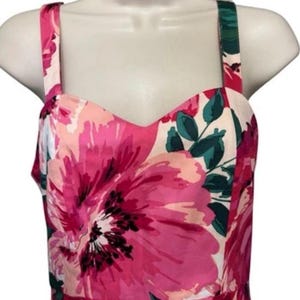 May include: A floral print dress with a sweetheart neckline and spaghetti straps. The dress features a vibrant pattern of large pink flowers with green leaves against a white background. The dress is displayed on a mannequin.