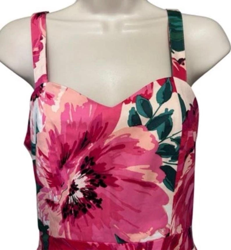 May include: A floral print dress with a sweetheart neckline and spaghetti straps. The dress features a vibrant pattern of large pink flowers with green leaves against a white background. The dress is displayed on a mannequin.