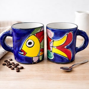 Może przedstawiać: Biały ceramiczny kubek z czarnym uchem i napisem "But First, Coffee" w czarnym kolorze. Rozmyte tło sugeruje przytulną atmosferę.