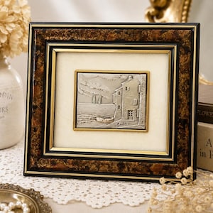 May include: A framed artwork featuring a detailed metal relief of a coastal scene with buildings, boats, and the sea. The frame has a dark brown, textured outer border, a black inner border, and a gold inner frame. The artwork is set against a cream-colored background.