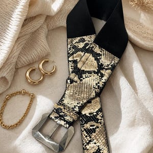 Puede incluir: Un cinturón negro con estampado de piel de serpiente y hebilla plateada. El cinturón tiene una banda elástica ancha y un estampado de piel de serpiente en tonos beige, marrón y negro. También se ven pendientes de aro dorados y una pulsera dorada.