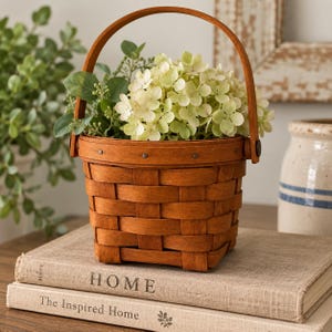 May include: A round, woven wooden basket with a fixed arched handle, filled with light green hydrangea flowers and foliage. The basket features a horizontal band secured with small metal rivets around the top rim, showcasing a natural wood grain.