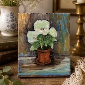 May include: An oil painting on canvas depicting a potted plant with three large, pale white flowers and green leaves. The plant sits in a terracotta-colored pot on a flat surface. The background features textured, dark brown and blue brushstrokes.