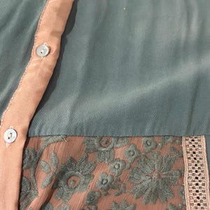 Op de afbeelding: Close-up van een teal-kleurige blouse met perzikkleurige accenten. De blouse heeft een perzikkleurige knoopsluiting en een bloemen kanten paneel. De halslijn en manchetten hebben een delicate, patroonafwerking. De stof lijkt een zacht, vloeiend materiaal te zijn.