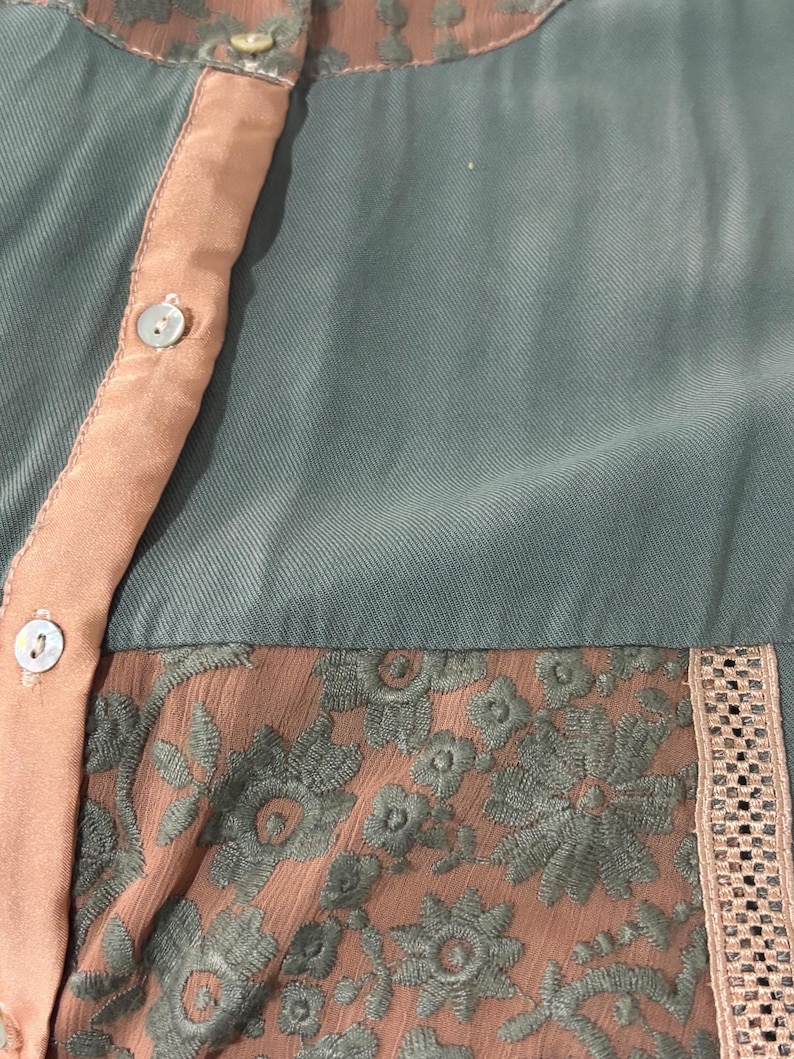 Op de afbeelding: Close-up van een teal-kleurige blouse met perzikkleurige accenten. De blouse heeft een perzikkleurige knoopsluiting en een bloemen kanten paneel. De halslijn en manchetten hebben een delicate, patroonafwerking. De stof lijkt een zacht, vloeiend materiaal te zijn.