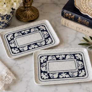 May include: Two rectangular ceramic trays featuring a cream-colored base with a distinct crackle glaze finish. Each tray displays a dark blue border pattern consisting of stylized rabbits and foliage surrounding a central blank rectangular area.