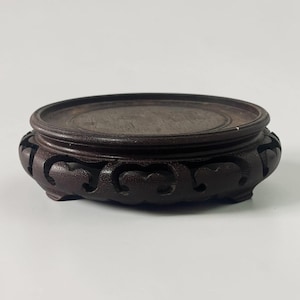 May include: A dark brown, circular wooden display stand featuring a flat top surface and a carved, openwork base with repeating cloud-like patterns. The stand is supported by small, integrated feet, showcasing a smooth, polished finish.