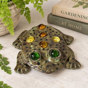 May include: A ceramic frog figurine with a mottled brown and green glaze, featuring two large, round, vibrant green glass eyes and four smaller, translucent amber and yellow glass cabochons embedded along its back in a symmetrical pattern.