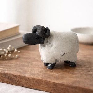 Könnte beinhalten: Keramik-Schaf-Figur mit schwarzem Kopf und Hörnern sowie weißem Körper. Das Schaf steht auf einer Holzoberfläche. Die Figur ist ein dekorativer Artikel.