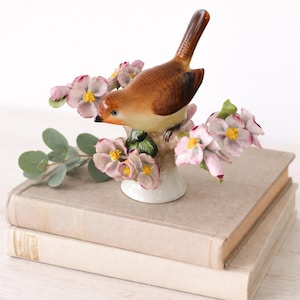 May include: A ceramic bird figurine perched on a flowering branch, with pink and yellow blossoms. The bird has brown and tan feathers and blue eyes. The figurine sits atop two stacked books with beige covers.