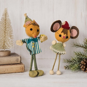 May include: Two felt mouse ornaments. The left mouse wears a green striped shirt with a bow tie and a green pointed hat. The right mouse wears a green dress with lace trim and a small red hat. Both have round ears, black eyes, and thin legs.