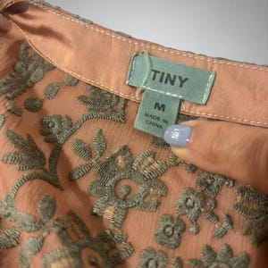 Op de afbeelding: Close-up van een perzikroze blouse met gedetailleerde grijze bloemenborduurwerk. De blouse heeft een delicate, transparante stof en een klein label met de tekst "TINY" en "M Made in China". Het borduurwerk voegt een gestructureerd, elegant detail toe.