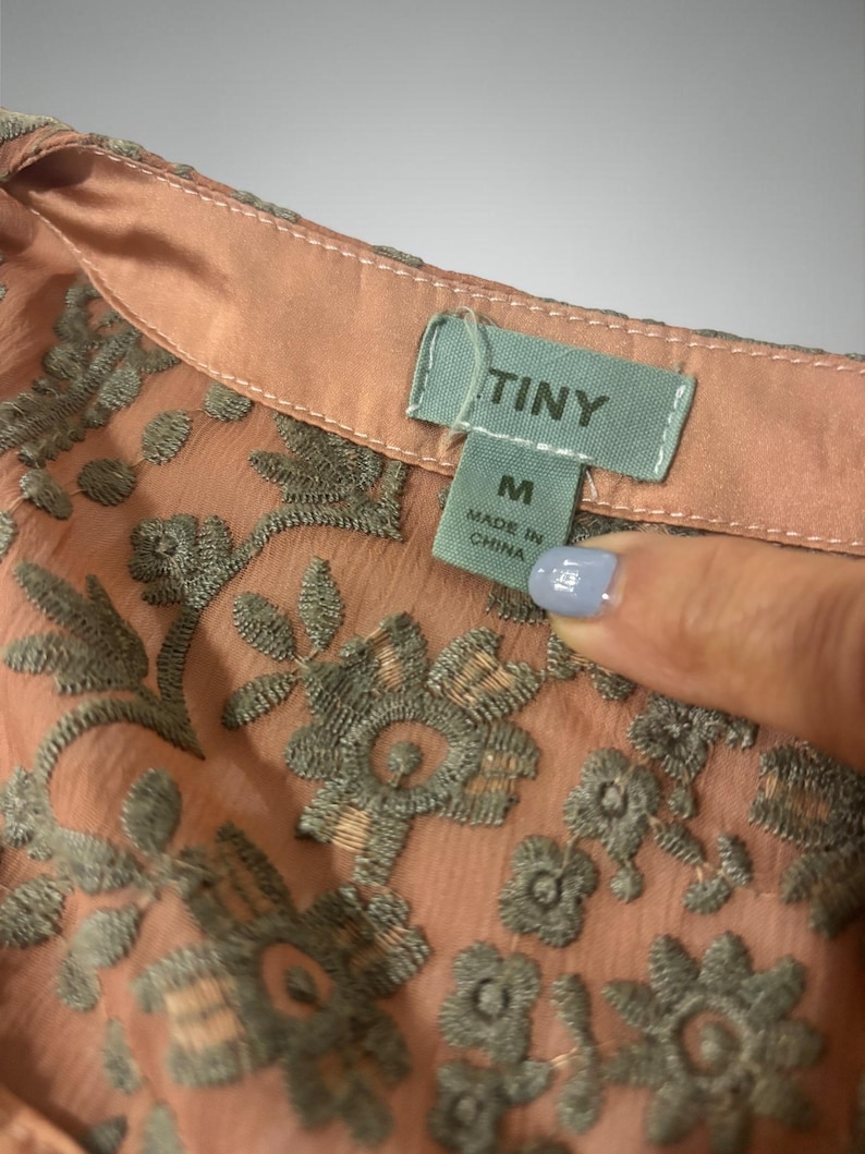 Op de afbeelding: Close-up van een perzikroze blouse met gedetailleerde grijze bloemenborduurwerk. De blouse heeft een delicate, transparante stof en een klein label met de tekst "TINY" en "M Made in China". Het borduurwerk voegt een gestructureerd, elegant detail toe.