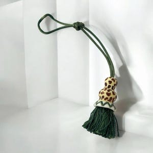 May include: A decorative tassel with a green cord loop. The tassel features a cream-colored, bulbous ceramic bead with brown spots, a white and green patterned base, and a dark green fringe.