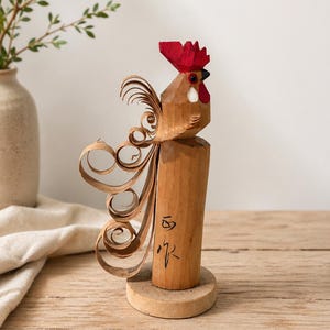Könnte beinhalten: Holzhahn-Skulptur mit rotem Kamm und weißer Wamme. Der Hahn sitzt auf einem Holzsockel, mit einem dekorativen Schwanz aus gelockten Holzspänen. Die Skulptur hat japanische Schriftzeichen auf der Seite.