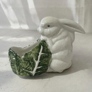 Könnte beinhalten: Eine weiße Keramik-Hasenfigur mit einem grünen, blattförmigen Pflanzgefäß. Der Hase sitzt aufrecht, mit seinen Pfoten auf dem Blatt. Der Pflanztopf ist tiefgrün mit weißen Adern.