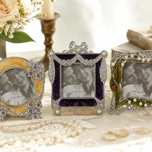 May include: Three decorative photo frames in silver, yellow, and purple. The frames have ornate designs with floral and leaf accents. The frames are arranged on a lace tablecloth with pearls and flower petals.