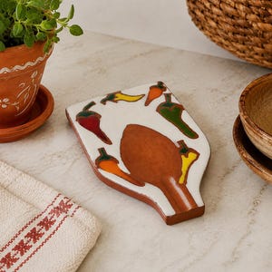 Puede incluir: Un reposacucharas de cerámica con esmalte blanco, una zona central de color terracota marrón y motivos de chiles pintados a mano en rojo, naranja, amarillo y verde. El reposacucharas tiene una forma cónica con un cuenco ancho y un mango estrecho.