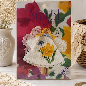 May include: A Counted Cross Stitch Angel Doll craft kit package featuring a yellow-haired angel figure with white lace wings and a dress embroidered with a yellow jonquil flower and a small blue butterfly, set against a floral background.