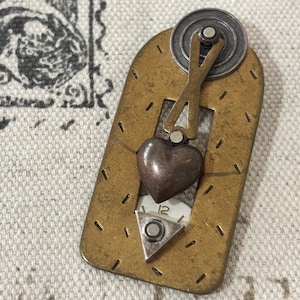 May include: A steampunk-style brooch with a brass-colored rectangular base, featuring a heart-shaped charm and a small clock face. The brooch has a silver-colored gear and a mechanical design, with a vintage aesthetic.