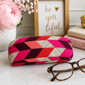 Könnte beinhalten: Ein Brillenetui mit geometrischem Muster in Rosa-, Rot- und Burgundertönen liegt neben einer Brille mit Schildpattrahmen. Eine goldene Vogelfigur und ein gerahmter Druck mit der Aufschrift "be you tiful" befinden sich im Hintergrund.