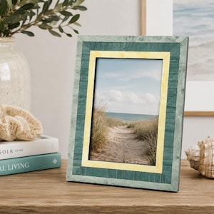 May include: A rectangular photo frame with a teal and yellow border. The frame holds a picture of a sandy path leading to the ocean. The frame is displayed on a wooden surface, next to a seashell and books.