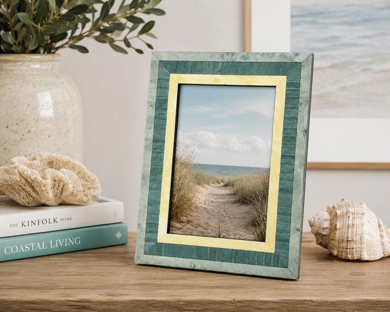 May include: A rectangular photo frame with a teal and yellow border. The frame holds a picture of a sandy path leading to the ocean. The frame is displayed on a wooden surface, next to a seashell and books.