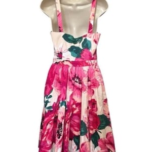 May include: A floral print dress with a fitted bodice and flared skirt. The dress features a pattern of large pink and magenta flowers with green leaves against a cream background. It has a scoop neckline and wide shoulder straps. The dress is sleeveless.
