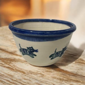 May include: A ceramic bowl with a white base and a blue rim and band. The bowl features hand-painted blue cat illustrations around the exterior. The bowl is sitting on a wooden surface.