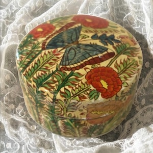 Puede incluir: Caja redonda decorativa con tapa, con un diseño pintado a mano. La caja muestra una mariposa con alas azules y bordes marrón-rojizos, rodeada de flores rojas y follaje verde. El fondo es de color amarillo claro.