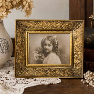 May include: A rectangular picture frame with an ornate, embossed gold finish featuring intricate scrollwork and leaf patterns. The frame holds a sepia-toned photograph of a young person with curly hair and a bow, holding a bouquet of flowers.