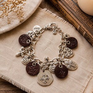 May include: Silver charm bracelet with various charms, including hearts, crosses, and floral designs. The bracelet also features brown leather-like circular charms. The word "Amor" is on one of the charms.