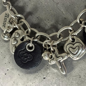 May include: Silver charm bracelet with multiple charms, including hearts, a cross, a bell, and a black leather charm with a floral design. One charm has the word "Amor" engraved.