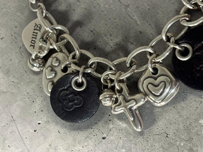 May include: Silver charm bracelet with multiple charms, including hearts, a cross, a bell, and a black leather charm with a floral design. One charm has the word "Amor" engraved.
