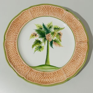 May include: A round decorative plate with a palm tree design. The center of the plate features a green and brown palm tree on a white background. The rim has a textured, woven pattern in tan and is bordered by a green edge.