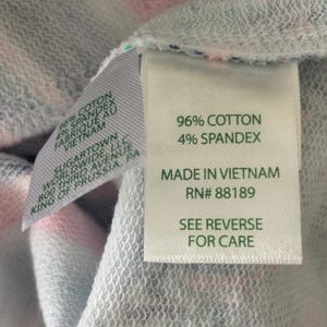 May include: Close-up of a garment tag with the text "96% COTTON 4% SPANDEX MADE IN VIETNAM RN# 88189 SEE REVERSE FOR CARE". The fabric is a mix of pink, white, and blue.