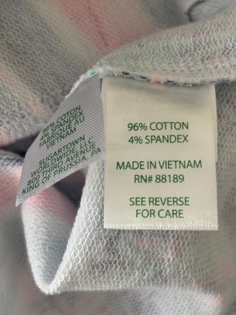 May include: Close-up of a garment tag with the text "96% COTTON 4% SPANDEX MADE IN VIETNAM RN# 88189 SEE REVERSE FOR CARE". The fabric is a mix of pink, white, and blue.