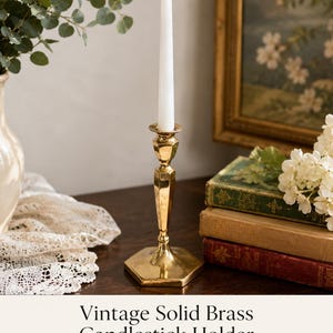 May include: A polished brass candlestick holder featuring a hexagonal base and a slender, faceted stem. The holder supports a single white taper candle with a lit flame. The metal surface is reflective and smooth, showcasing a metallic gold finish.