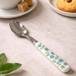 May include: A stainless steel spoon with a white handle decorated with blue floral patterns. The spoon is placed on a light-coloured surface, with a cup of coffee, sugar cubes, and biscuits in the background.