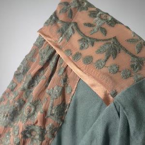 Op de afbeelding: Close-up van een kledingstuk met een combinatie van texturen en kleuren. Het ontwerp omvat een gedeelte van stoffig roze stof met geborduurde bloempatronen in gedempt groen. De hoofdbody van het kledingstuk is een zachte, gedempte teal stof.