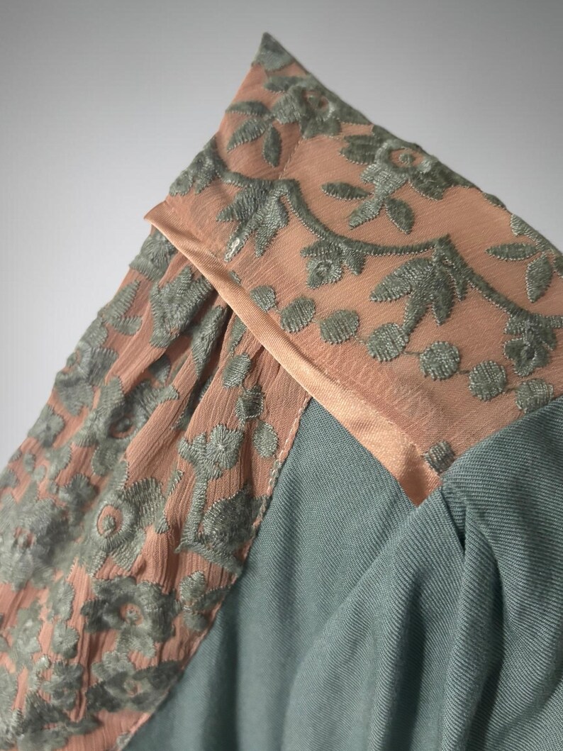 Op de afbeelding: Close-up van een kledingstuk met een combinatie van texturen en kleuren. Het ontwerp omvat een gedeelte van stoffig roze stof met geborduurde bloempatronen in gedempt groen. De hoofdbody van het kledingstuk is een zachte, gedempte teal stof.