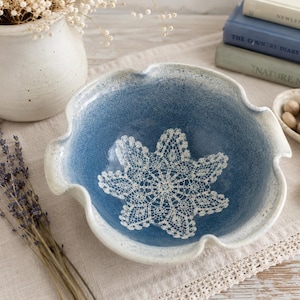 May include: A ceramic bowl with a scalloped rim, featuring a deep blue speckled interior and a white exterior. The center of the bowl displays a white, intricate lace-like doily pattern imprinted into the surface, creating a textured design.