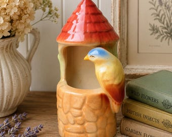 May include: A ceramic birdhouse figurine featuring a red conical roof and a tan stone-textured base. A small, sculpted bird with a blue head, yellow body, and orange-tipped wings is perched on the side of the opening of the structure.