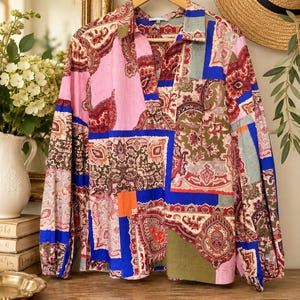 May include: A long-sleeve button-down shirt featuring a vibrant patchwork design with paisley patterns in shades of pink, blue, olive green, and orange. The shirt has a classic collar, a V-neckline, and cuffed sleeves, displayed on a wooden hanger.