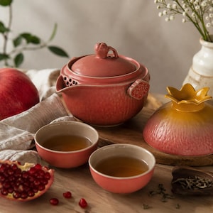 Può includere: Un set da tè in ceramica composto da una teiera color corallo testurizzata con coperchio abbinato, due piccole ciotole riempite di tè e una ciotola separata con un coperchio a forma di melograno giallo e rosa, con un melograno fresco.